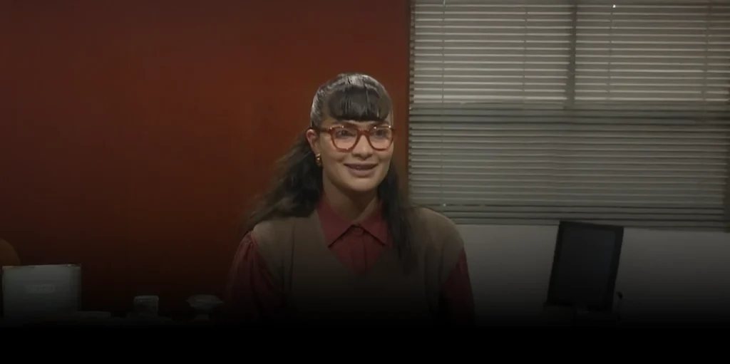 Mujer joven, con gafas de marco grueso de color naranja y un flequillo recto, viste un chaleco marrón sobre una camisa roja.