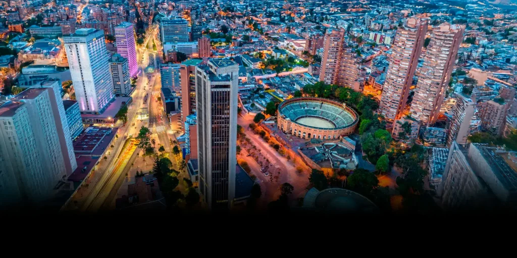 Vista nocturna aérea de Bogotá mostrando rascacielos iluminados, la Plaza de Toros La Santamaría circular y el denso paisaje urbano con luces de la ciudad
